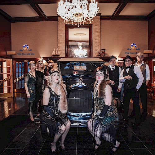 A group of people in 1920s attire pose in front of a vintage car inside a luxurious, chandelier-lit room, creating a glamorous setting.