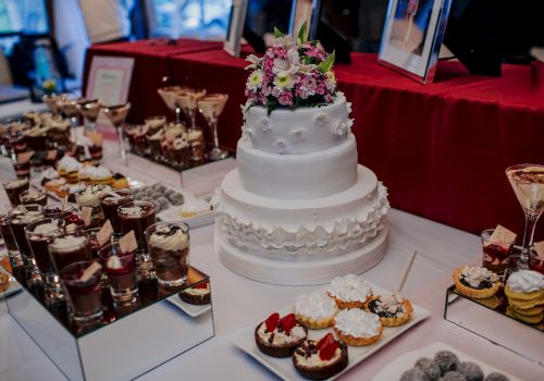 The image shows a dessert table with a tiered white cake adorned with flowers, surrounded by various pastries, sweets, and dessert cups.