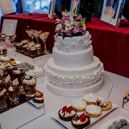 The image shows a dessert table with a tiered white cake adorned with flowers, surrounded by various pastries, sweets, and dessert cups.