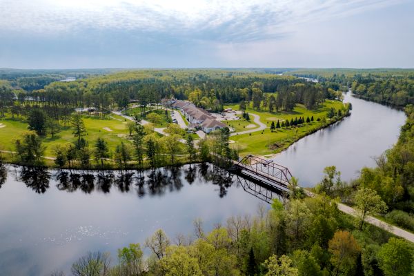 An aerial view of a serene landscape featuring a river, a bridge, buildings, and lush greenery, under a partly cloudy sky.