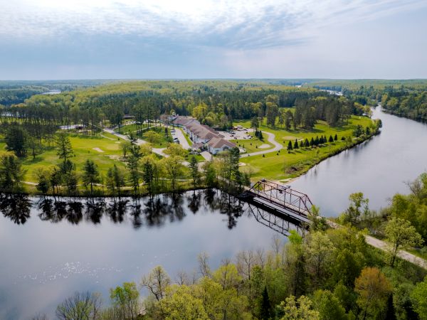An aerial view of a serene landscape featuring a river, a bridge, buildings, and lush greenery, under a partly cloudy sky.