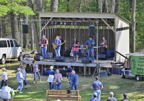 A band performing on an outdoor stage surrounded by a forest, with an audience watching and dancing on the grass.