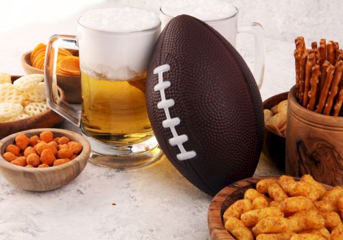 The image features a football, beer mugs, and various snacks including pretzels, cheese balls, and crackers, set on a table.