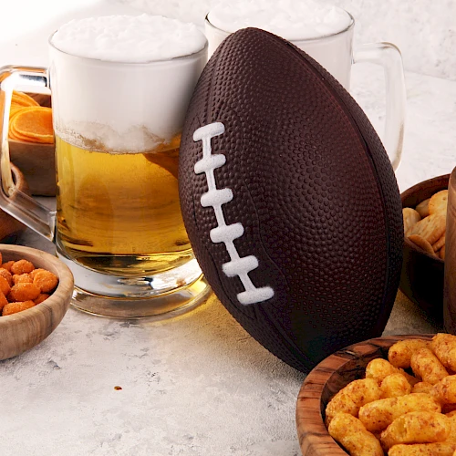 The image features a football, beer mugs, and various snacks including pretzels, cheese balls, and crackers, set on a table.