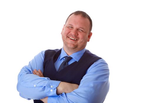 The image shows a smiling man in a blue shirt and dark vest, with his arms crossed, against a plain white background.