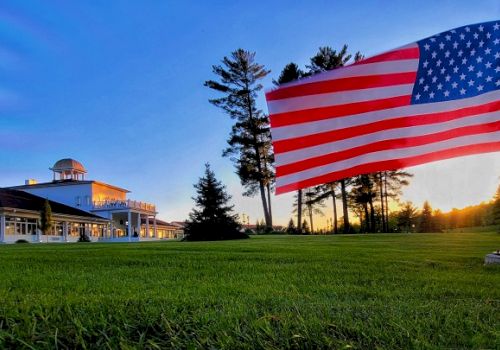 An American flag stands in the foreground with a large building and trees in the background during sunset.