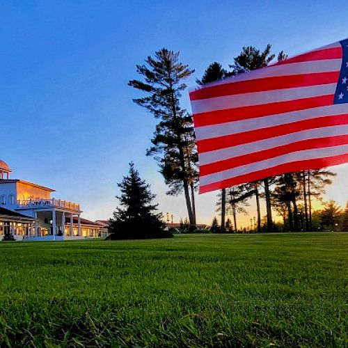 An American flag stands in the foreground with a large building and trees in the background during sunset.