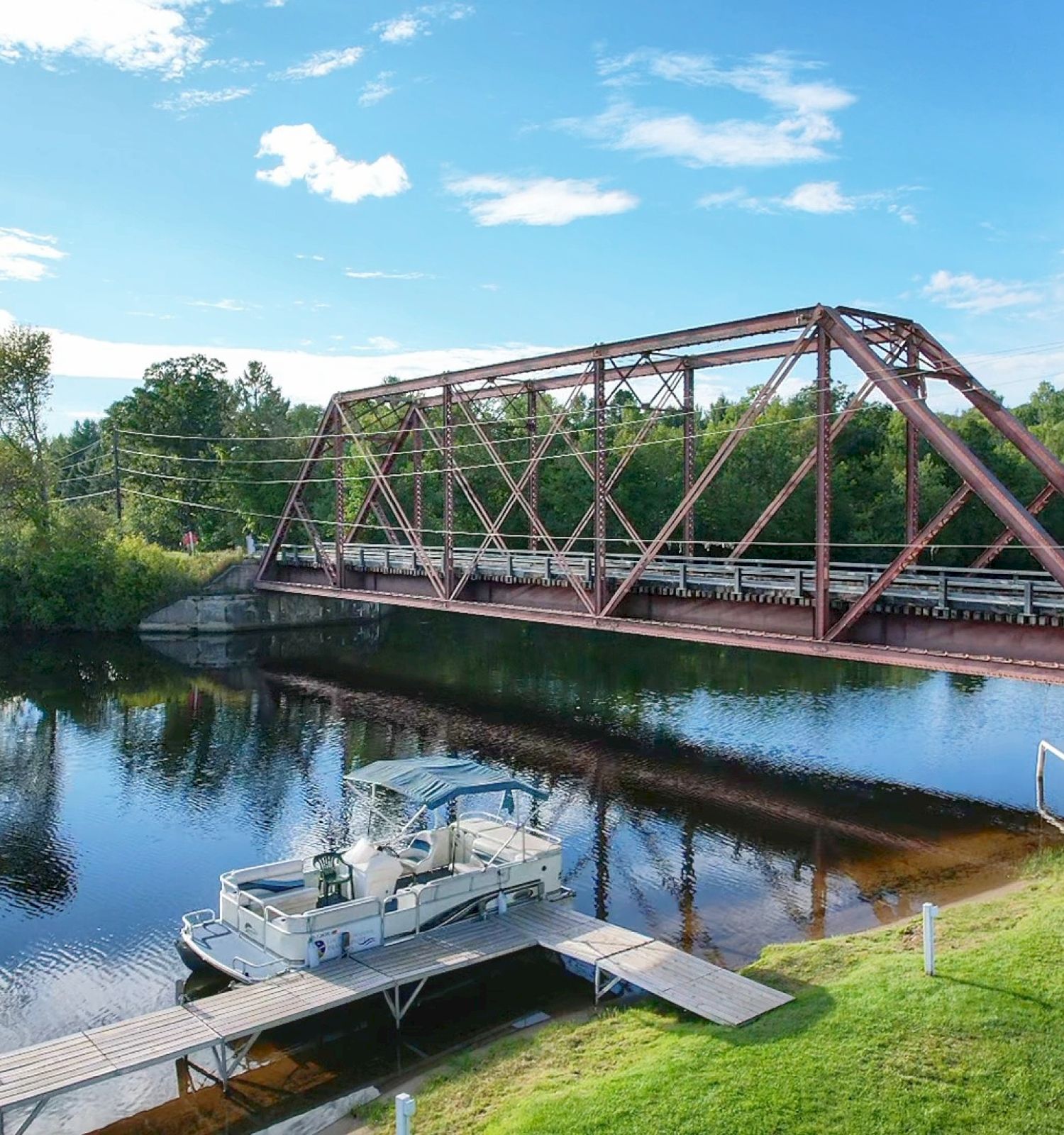 A boat docked near a river with a red truss bridge in the background, surrounded by trees and a clear blue sky ending the sentence.