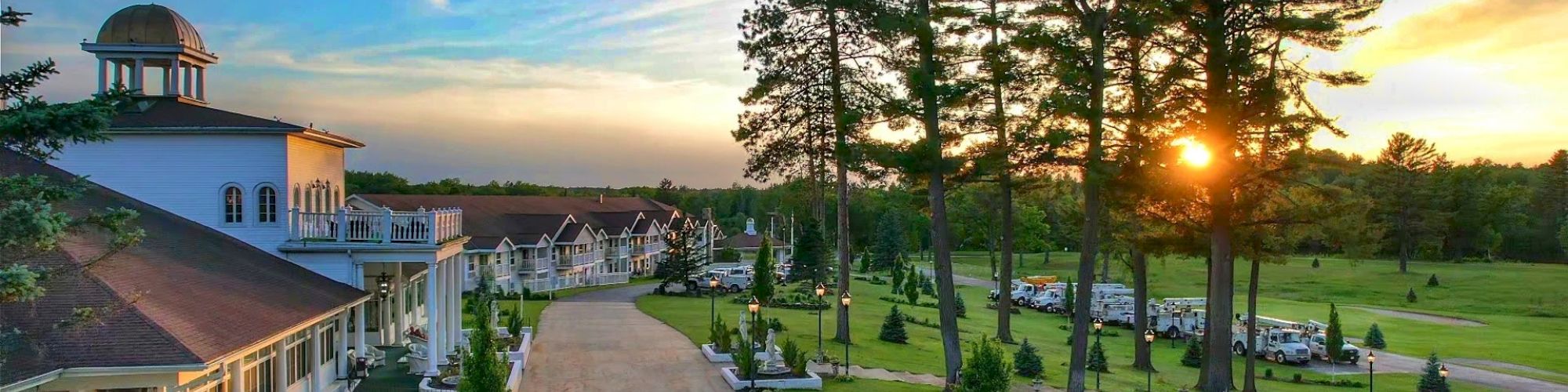The image depicts a scenic landscape of a large building with a driveway, lush green lawns, tall trees, and a sunset in the background.