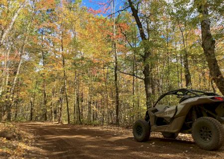A off-road vehicle is parked on a dirt path surrounded by trees with autumn foliage under a clear blue sky.