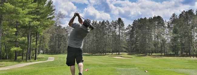 A person in a gray shirt and black shorts is playing golf on a green course surrounded by trees under a partly cloudy sky.