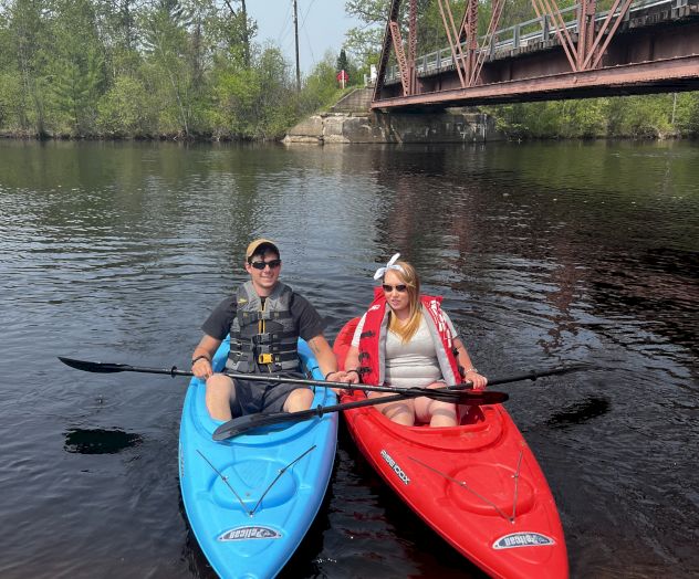 Two people in life jackets are sitting in blue and red kayaks on a calm river, with a bridge and trees in the background.