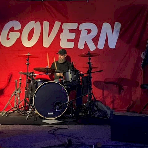 Three people are performing on stage with musical instruments in front of a red backdrop that says "GOVERN".