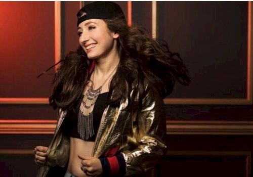 A person smiling, wearing a black cap backwards, a gold and black jacket, layered necklaces, and a crop top, standing against a wall with geometric accents.