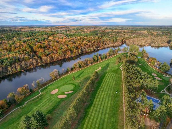 An aerial view of a golf course surrounded by trees, a winding river, and a tennis court, set in a colorful autumn landscape.