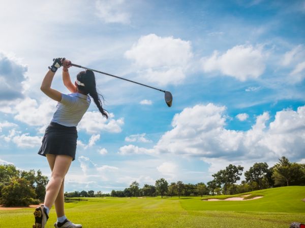 A person is playing golf, swinging a club on a lush green course under a blue sky with scattered clouds.