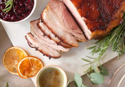 A sliced roasted meat dish with herbs, a bowl of sauce, dried orange slices, and a bowl of berry sauce are displayed on a white surface.