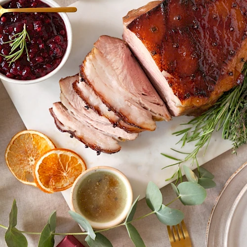 A sliced roasted meat dish with herbs, a bowl of sauce, dried orange slices, and a bowl of berry sauce are displayed on a white surface.