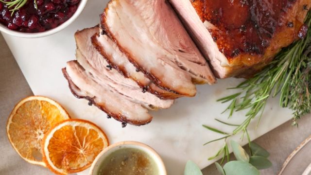 A sliced roasted meat dish with herbs, a bowl of sauce, dried orange slices, and a bowl of berry sauce are displayed on a white surface.