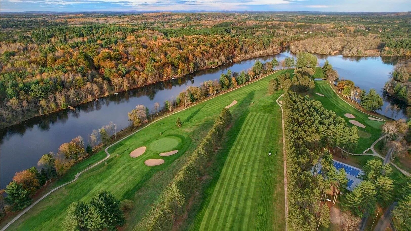 Aerial view of a golf course with vibrant green fairways, surrounded by a river and lush trees under a blue sky with scattered clouds.
