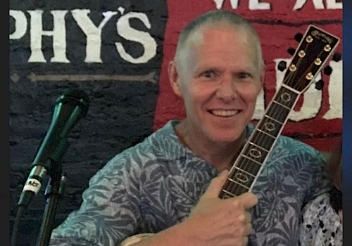 A person is holding an acoustic guitar and standing in front of a microphone with a wall featuring partial text in the background.