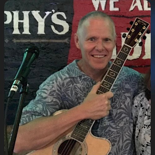 A person is holding an acoustic guitar and standing in front of a microphone with a wall featuring partial text in the background.