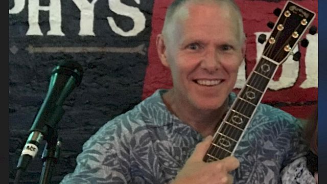 A person is holding an acoustic guitar and standing in front of a microphone with a wall featuring partial text in the background.