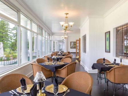 The image shows a sunlit dining area with large windows, wicker chairs, and set tables, featuring a garden view outside.