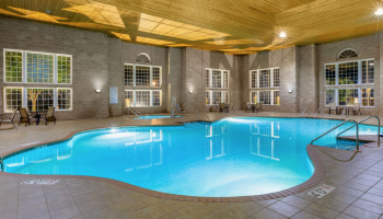 An indoor swimming pool is illuminated with blue water. There are lounge chairs and tables on the perimeter, with large windows and a wood ceiling.